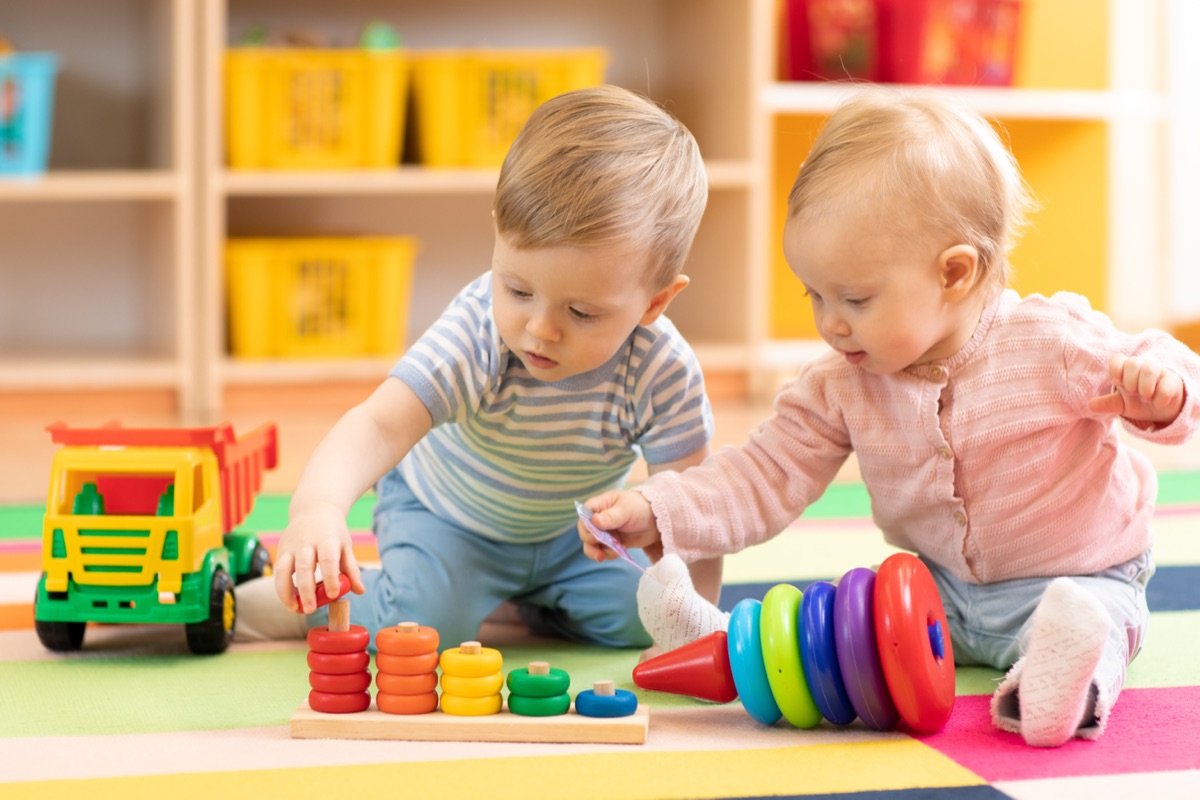 Two toddlers playing with stacking rings and a toy truck on a colourful play mat